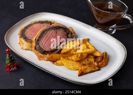 Wellington di manzo con fette di patate al forno e salsa demi-glace. Tradizionale piatto festivo di filetto di manzo con funghi e prosciutto di Parma cotto in pasta sfoglia Foto Stock