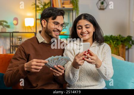 Felice è uscita dalla coppia indiana che ha in mano soldi contanti e pianificazione smartphone discutendo di investimenti insieme seduti sul divano nel soggiorno di casa. Famiglia sorridente di successo dopo aver vinto il jackpot della lotteria Foto Stock