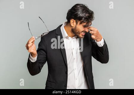 Stanco uomo d'affari indiano si toglie gli occhiali, sente dolore agli occhi, è troppo lavoro bruciato da lunghe ore di lavoro. Sonnolento e esausto ragazzo arabo che sfrega gli occhi isolato su sfondo grigio studio Foto Stock