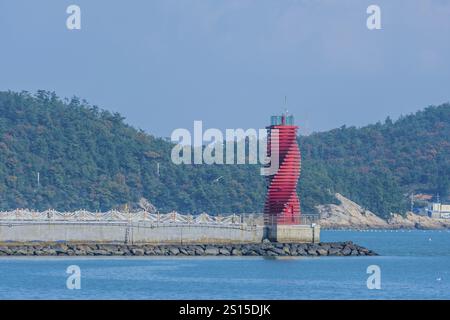Un faro rosso unico sorge sulla costa rocciosa con uno sfondo di foresta contro l'oceano, situato a Wando, Corea del Sud, Asia Foto Stock