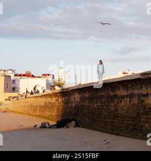 Passeggiate turistiche femminili sulle mura della città con la storica Medina alle spalle e la costa rocciosa lasciata nella città di Essaouira, 31 dicembre 2024 Foto Stock