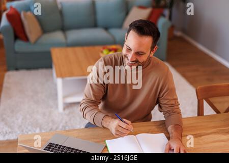 Vista ad angolo alto dell'imprenditore che utilizza il laptop e scrive in blocco note mentre lavora in ufficio domestico Foto Stock