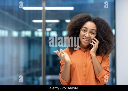 Ritratto di una gioiosa donna d'affari, lavoratrice d'ufficio con un telefono in mano che parla con i clienti, sorride e guarda la telecamera, in piedi vicino alla finestra di un edificio di uffici sul suo posto di lavoro. Foto Stock