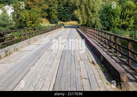Vecchio ponte di legno con ringhiere su un fiume circondato da lussureggianti alberi verdi. Fotografia prospettica di un paesaggio rurale. Foto Stock