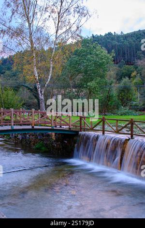 Una vista panoramica di una piccola cascata sul fiume Saja a Cabezon de la Sal, Cantabria, Spagna. L'immagine presenta un affascinante ponte di legno che attraversa Foto Stock