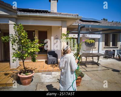 Una donna innaffia le piante in un giardino soleggiato con pannelli solari sul tetto. La scena cattura un momento di cura e sostenibilità, ambientato in Foto Stock