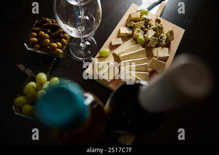 Una saporita disposizione di vari formaggi, uve fresche, olive e bottiglie di vino, perfetta per una celebrazione elegante o una delizia culinaria. Foto Stock