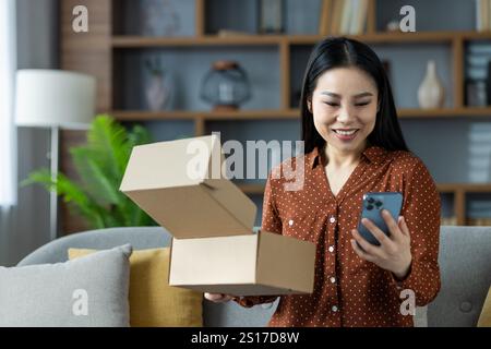 Donna asiatica a casa che tiene il pacchetto e controlla i dettagli sullo smartphone, esprimendo gioia e soddisfazione. Concetto di shopping online, consegna, comodità e stile di vita moderno. Foto Stock
