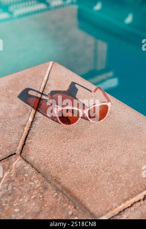 Occhiali da sole da donna con perle rosa sul ponte della piscina a bordo piscina con superficie in pietra ruvida. Piscina blu turchese. Gli occhiali da sole gettano un'ombra. Foto Stock