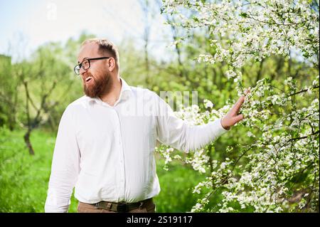 L'uomo allergico soffre di allergia stagionale in primavera nel giardino fiorito in primavera. Uomo barbuto con occhiali che allontanano il ramo di albero fiorito con fiori. Concetto di allergia primaverile Foto Stock