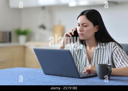Donna asiatica arrabbiata che parla al telefono controllando il portatile in cucina a casa Foto Stock