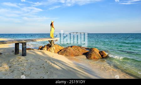 Un momento sereno si dispiega mentre una donna si erge su un molo di legno, osservando le tranquille acque dell'isola di Koh Munnork durante il tramonto. La luce dorata bagna il paesaggio, esaltando la bellezza della spiaggia. Foto Stock