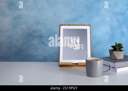 Nuove tendenze. Tavoletta moderna, tazza di caffè, libri e succulento tavolo bianco vicino alla parete blu Foto Stock