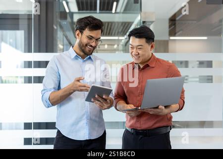 Due colleghi programmatori in abiti casual che discutono di un nuovo progetto software, uomini in piedi alla finestra con un portatile, sorridenti, che parlano. Foto Stock