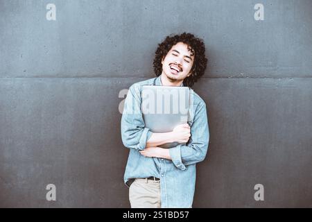 Un giovane uomo sorridente con capelli ricci che indossa una camicia in denim regge saldamente un notebook appoggiandosi su una parete grigia, esprimendo felicità e. Foto Stock