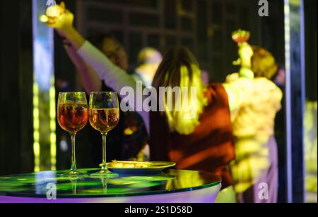 Festa colorata con bicchieri da vino in primo piano, amici che ballano e festeggiano con un'illuminazione vivace. Foto Stock