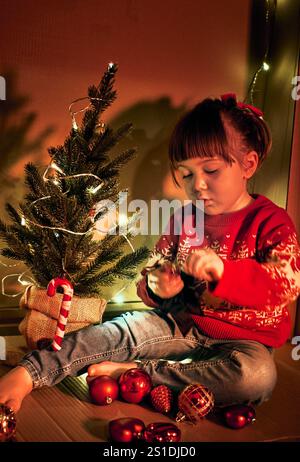 Preparazione per le vacanze e decorazioni natalizie. Una ragazza con un maglione rosso si siede accanto a un piccolo albero di Natale decorato, che regge un ornamento. Bambini in un ambiente accogliente Foto Stock