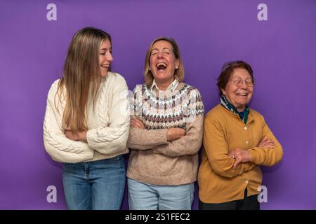 Nonna, madre e figlia che ridono con le braccia incrociate, in piedi fianco a fianco su sfondo viola, rappresentando tre generazioni di donne e t Foto Stock