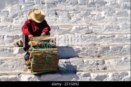 Venditore di legna da ardere Chiesa Capilla del Calvario Chichicastenango Guatemala | CHICHICASTENANGO, Guatemala — Un uomo che vende legna da ardere si riposa sui gradini della Capilla del Calvario, una chiesa imbiancata situata nel centro di Chichicastenango direttamente di fronte alla Chiesa Santo Tomas. Il venditore, indossando un cappello di paglia e una camicia rossa, è seduto dietro un fascio di legna legato con una corda verde. Aspetta clienti in questa città indigena Maya nelle alture del Guatemala. Foto Stock