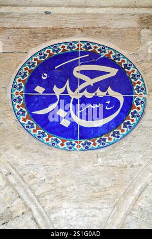 Moschea Rustem Pasha Iznik Tile Medallion Istanbul // ISTANBUL, Turchia — Un medaglione di ceramica con calligrafia araba che recita "Ya Sabur" (o paziente uno) decora la parete esterna della Moschea Rustem Pasha (nota anche come Rüstem Paşa Camii). L'iscrizione rappresenta uno dei 99 nomi di Dio nell'Islam, invocando la pazienza divina e la tolleranza. Questo elemento decorativo esemplifica il sofisticato lavoro di piastrelle di Iznik che copre la moschea, completato intorno al 1563 dal maestro architetto Mimar Sinan. La moschea ospita circa 2.300 piastrelle individuali in oltre 80 modelli distinti, che rappresentano Foto Stock