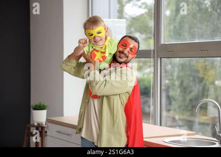 Padre felice con suo figlio piccolo vestito da supereroi che si diverte in cucina a casa Foto Stock