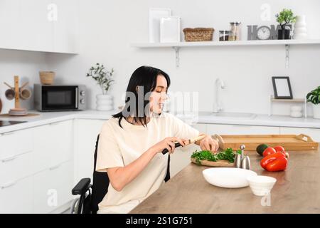 Bella giovane donna in sedia a rotelle che taglia verdure a casa Foto Stock