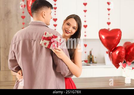 Giovane donna con regalo che abbraccia il suo ragazzo in cucina il giorno di San Valentino Foto Stock