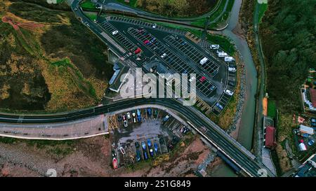 Vista aerea di un parcheggio costiero con auto, barche e un piccolo fiume. Strade e colline erbose circondano i parcheggi. Gli edifici sono visibili Foto Stock