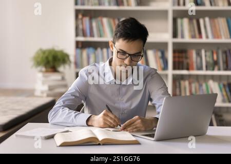 Un adolescente maschio concentrato in occhiali scrive lavori di carta in biblioteca Foto Stock