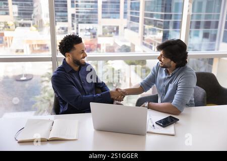 Due amici d'affari allegri e diversi che stringono la mano al notebook Foto Stock