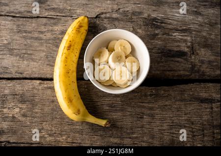 Vista dall'alto delle fette di banana fresche in una tazza accanto a una banana matura intera posizionata su tavole di legno rustiche. Foto Stock