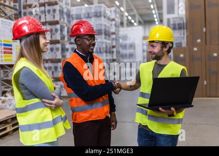 Tre operai edili che indossano attrezzature di sicurezza collaborano in un magazzino affollato, discutendo di logistica e lavoro di squadra. Due operai stringono la mano, indicando Foto Stock