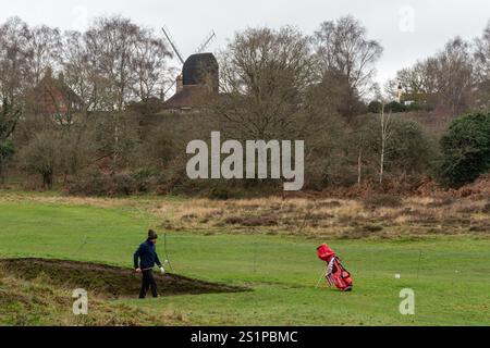 Golfista che gioca a golf al Reigate Heath Golf Club, Surrey, Inghilterra, Regno Unito, in inverno con il mulino a vento Reigate Heath sullo sfondo Foto Stock