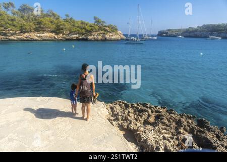 Madre e figlio in piedi su una scogliera, godendo di una vista mozzafiato delle barche a vela ancorate nelle acque turchesi di cala S'amarador, maiorca, spagna Foto Stock