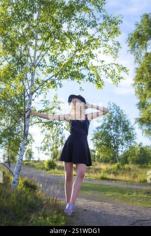 Giovane donna bionda in piedi accanto ad un albero di betulla in mezzo alla natura con un paesaggio in una serata estiva. Giovane donna bionda in piedi accanto a un albero in natura wi Foto Stock