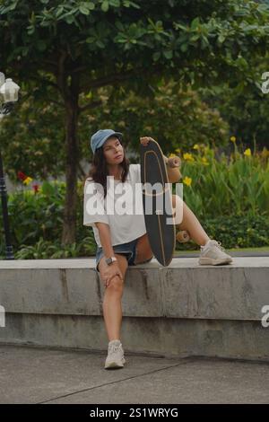 Una giovane donna siede comodamente su una parete bassa del parco tenendo in mano uno skateboard Foto Stock