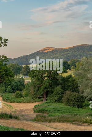 Caro percorso del castello di Eastnor, che si affaccia sul British Camp nelle Malvern Hills, immerso nella calda luce del sole con piccole nuvole sparse nel blu Foto Stock