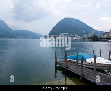 Vista di un molo con una barca su un lago in montagna e di un villaggio sull'altra sponda, vicino a Lugano nell'estate 2021 Foto Stock