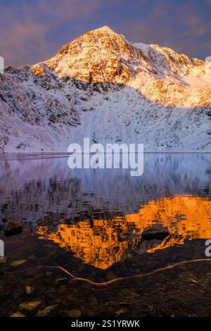 Una vista invernale all'alba di Yr Wyddfa, o Snowdon, riflessa nell'acqua ghiacciata di un Glaslyn sotto parzialmente congelato, Eryri (Snowdonia National Park), Galles Foto Stock