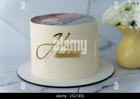 Torta di compleanno bianca minimalista con un lucido topper oro Happy Birthday sulla tavola, accompagnata da un vaso giallo su una superficie in marmo chiaro. Elegante cele Foto Stock