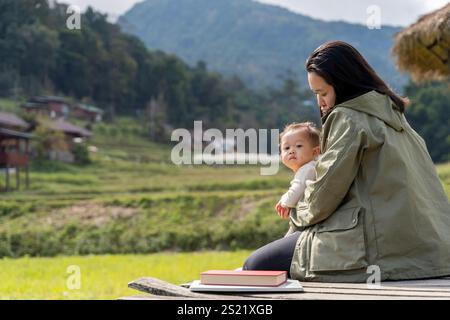 Vista posteriore di una donna seduta su un muro che abbraccia suo figlio. Mamma che bacia il suo bambino seduto sull'erba delle Alpi. Mote abbraccia sua figlia in un tramonto Foto Stock
