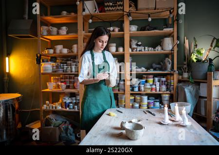 la vasaia femmina tiene e fissa la maniglia alla tazza di argilla per l'incollaggio del materiale, preparandosi per l'asciugatura e la cottura Foto Stock