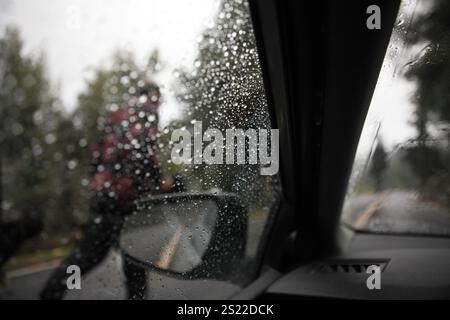 Uomo sinistro, non identificato, strada di campagna bagnata e diffusa, marciapiede, nebbia e alberi ad aghi visti attraverso gocce di pioggia sul parabrezza e sullo specchietto laterale dell'auto. Foto Stock