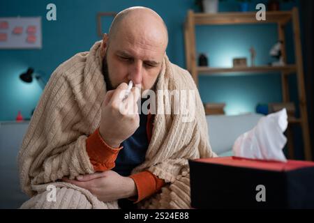 Un uomo malato di mezza età avvolto in una coperta usa uno spray nasale e sembra di avere un raffreddore Foto Stock