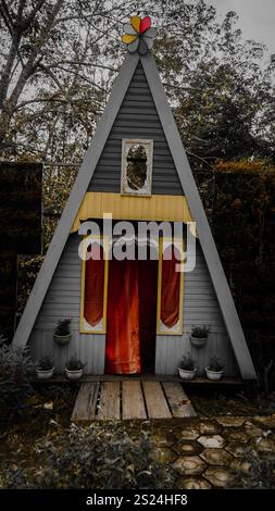 Una stravagante e triangolare sala giochi in legno con una porta a tenda rossa, circondata da piante e alberi. La casa dei giochi presenta un colorato motivo a rotellina Foto Stock