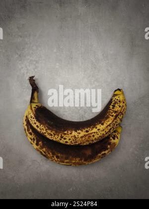 Due banane troppo mature maculate sono posizionate su una superficie grigia, che mostra la loro fase avanzata di maturazione Foto Stock