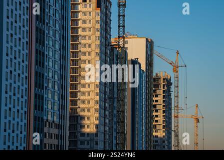 Gru da cantiere che operano su edifici residenziali moderni durante il tramonto, evidenziando lo sviluppo urbano e la crescita all'interno di un vivace paesaggio urbano Foto Stock