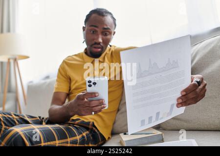 Un giovane afroamericano esamina i dati sul suo smartphone, completamente concentrato mentre è seduto a casa. Foto Stock