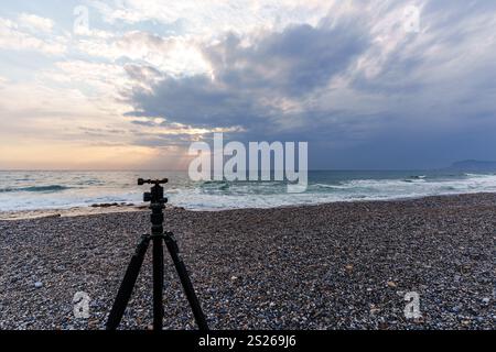 Una tranquilla spiaggia all'alba con un treppiede per fotocamera su una riva di ciottoli, che cattura le onde tranquille e il cielo esteso. Ideale per i concetti di fotografia Foto Stock