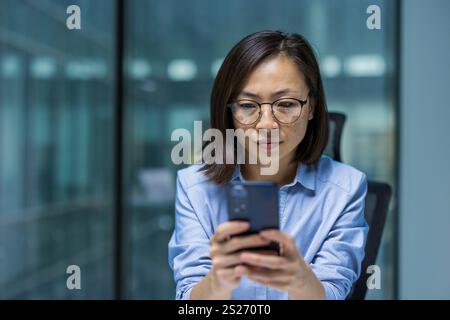 Donna seria e concentrata che utilizza l'app al telefono, donna d'affari asiatica che pensa a chiudere il messaggio sullo smartphone, che lavora all'interno dell'ufficio vicino alla finestra. Foto Stock